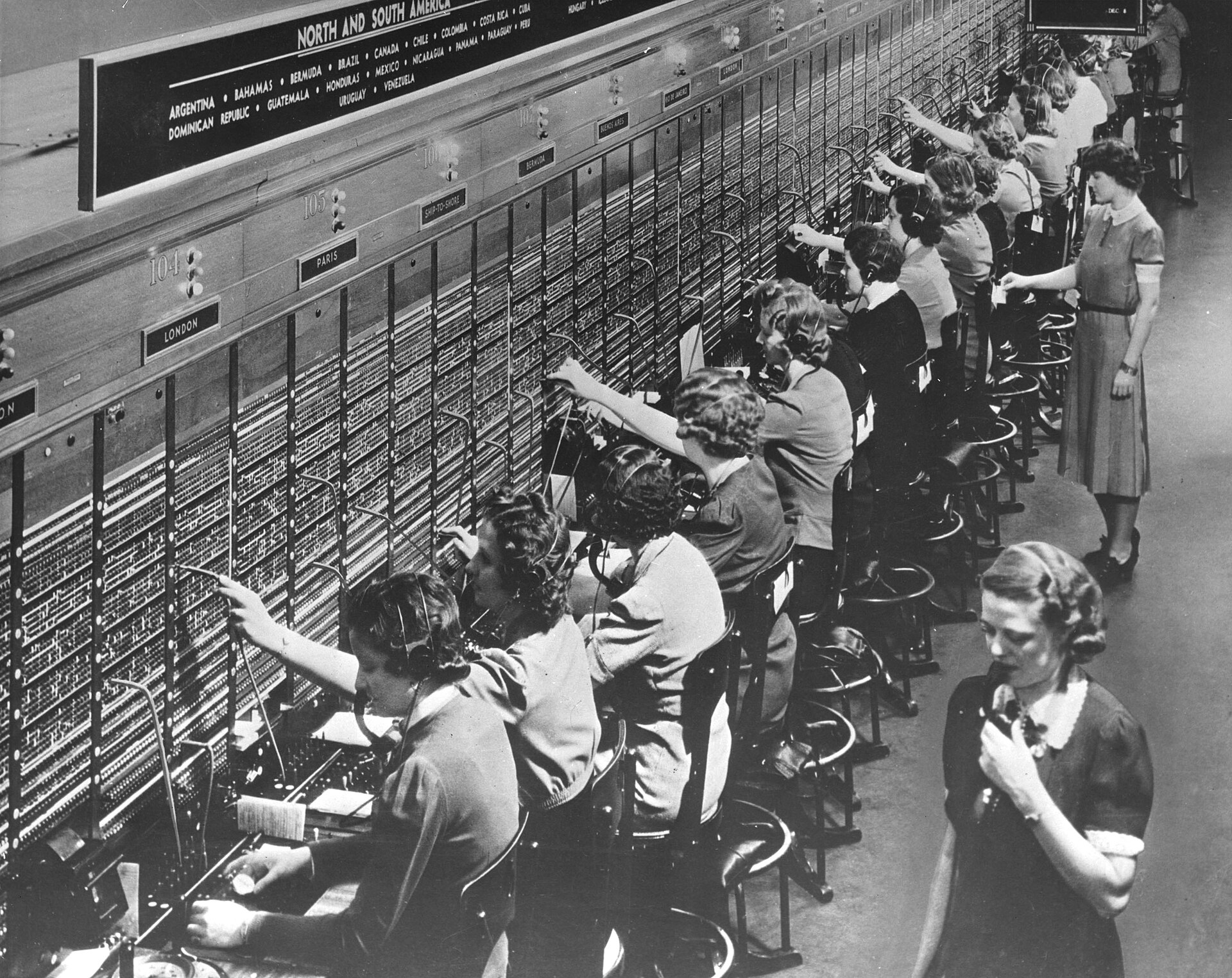 Rows of telephone operators in headsets at a long switchboard marked with destinations: London, Paris, Bermuda, ship-to-shore; a supervisor stands behind them.